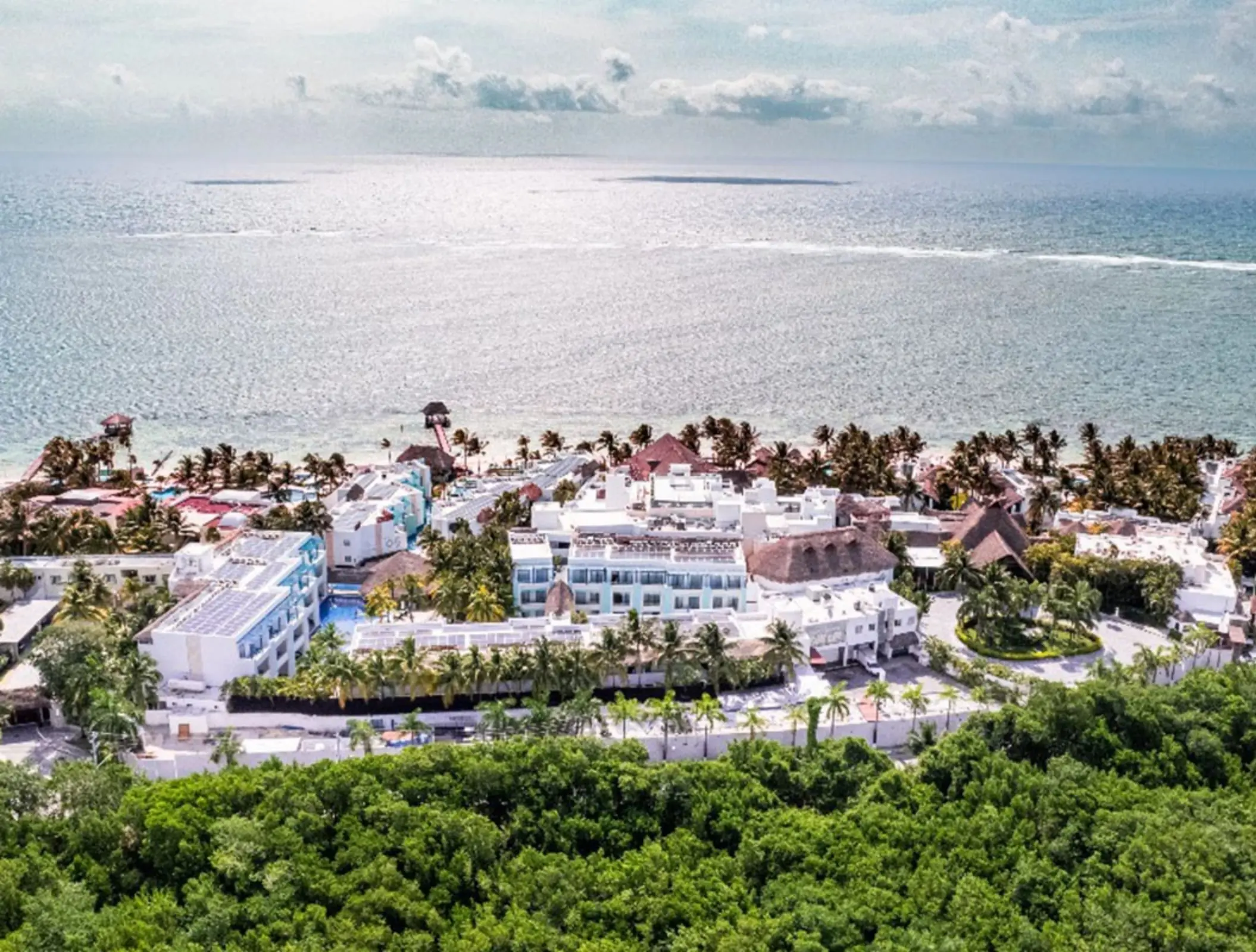 Aerial view of a beachfront resort with white buildings, lush palms, and the ocean stretching into the horizon.