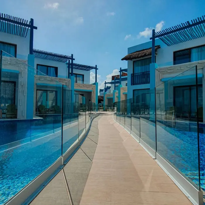 Swim-up suites lining a crystal-clear pool with a walkway, glass railing, and bright blue skies above.