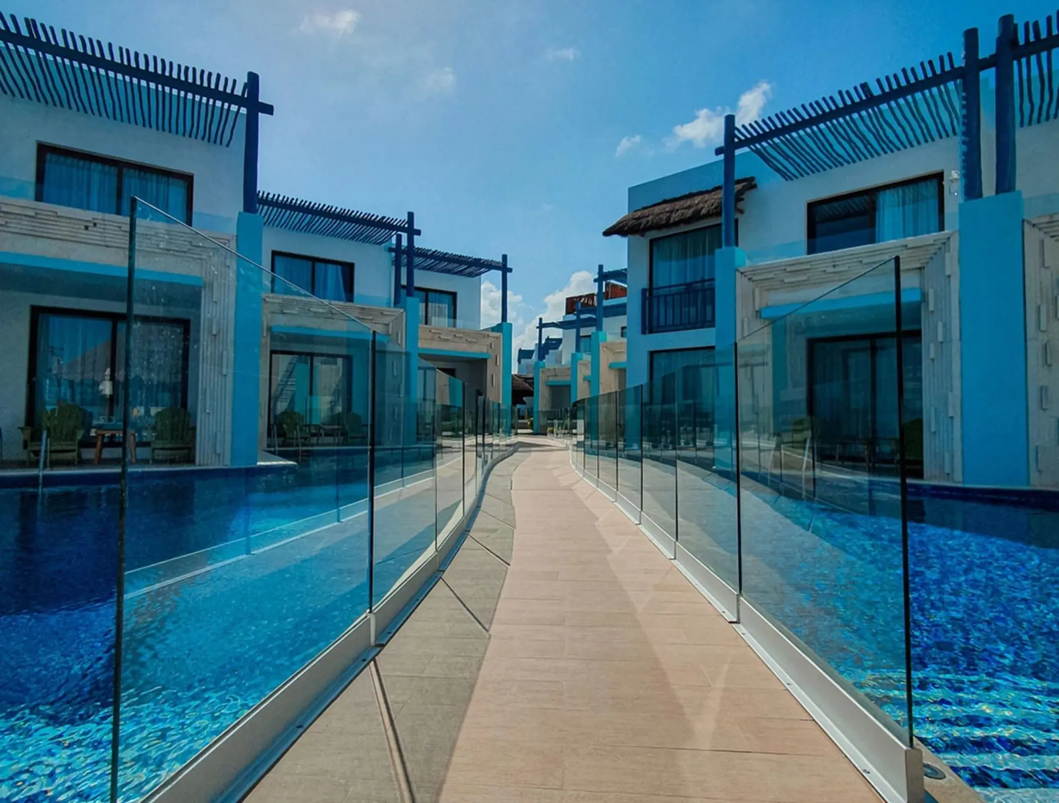 Swim-up suites lining a crystal-clear pool with a walkway, glass railing, and bright blue skies above.