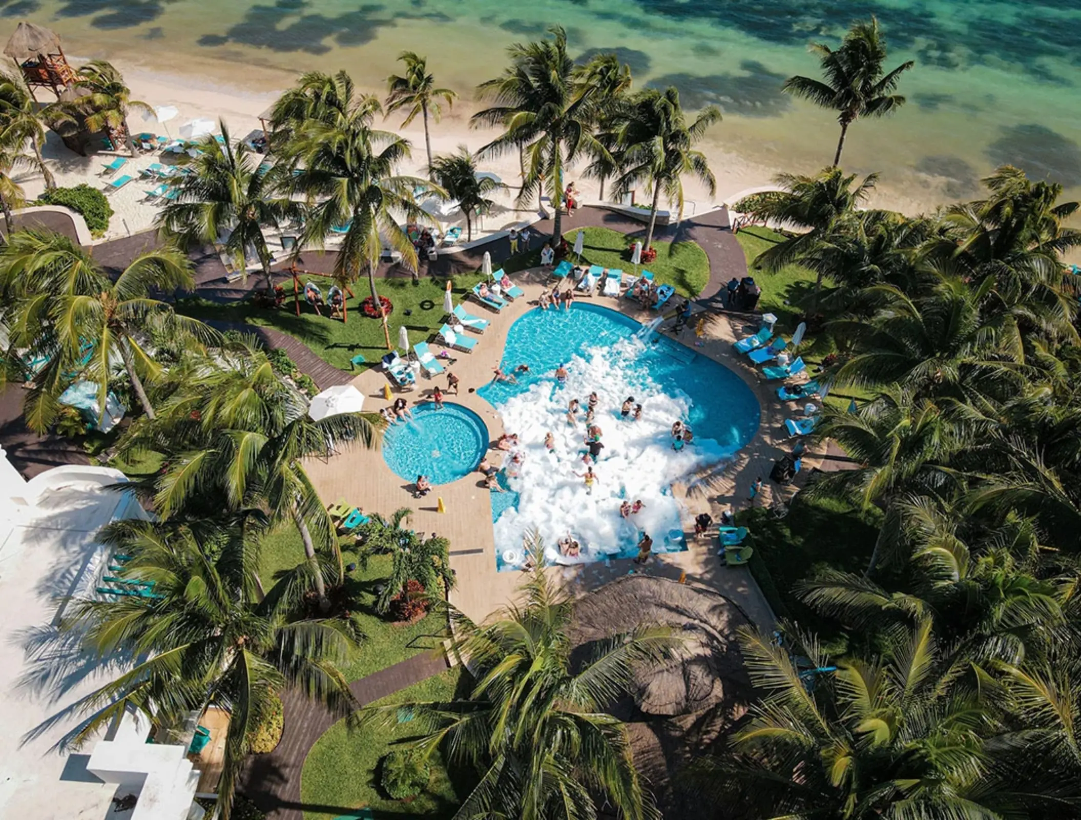 Foam party at a beachfront pool surrounded by palm trees and loungers, with guests enjoying the lively atmosphere.