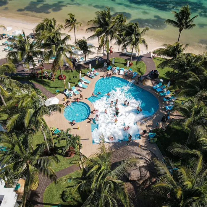 Foam party at a beachfront pool surrounded by palm trees and loungers, with guests enjoying the lively atmosphere.