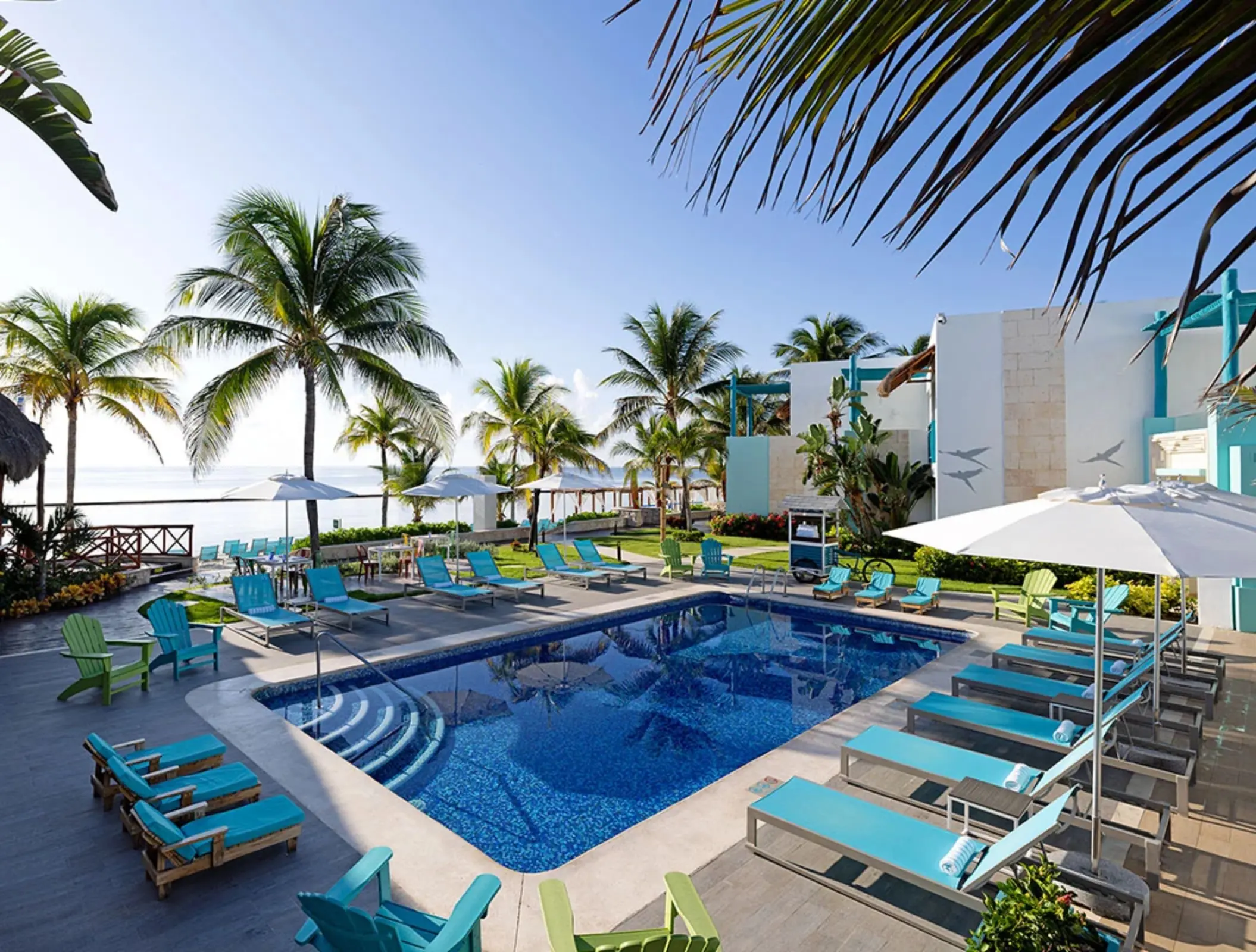 Tranquil beachfront pool with turquoise loungers, umbrellas, and palm trees nearby.