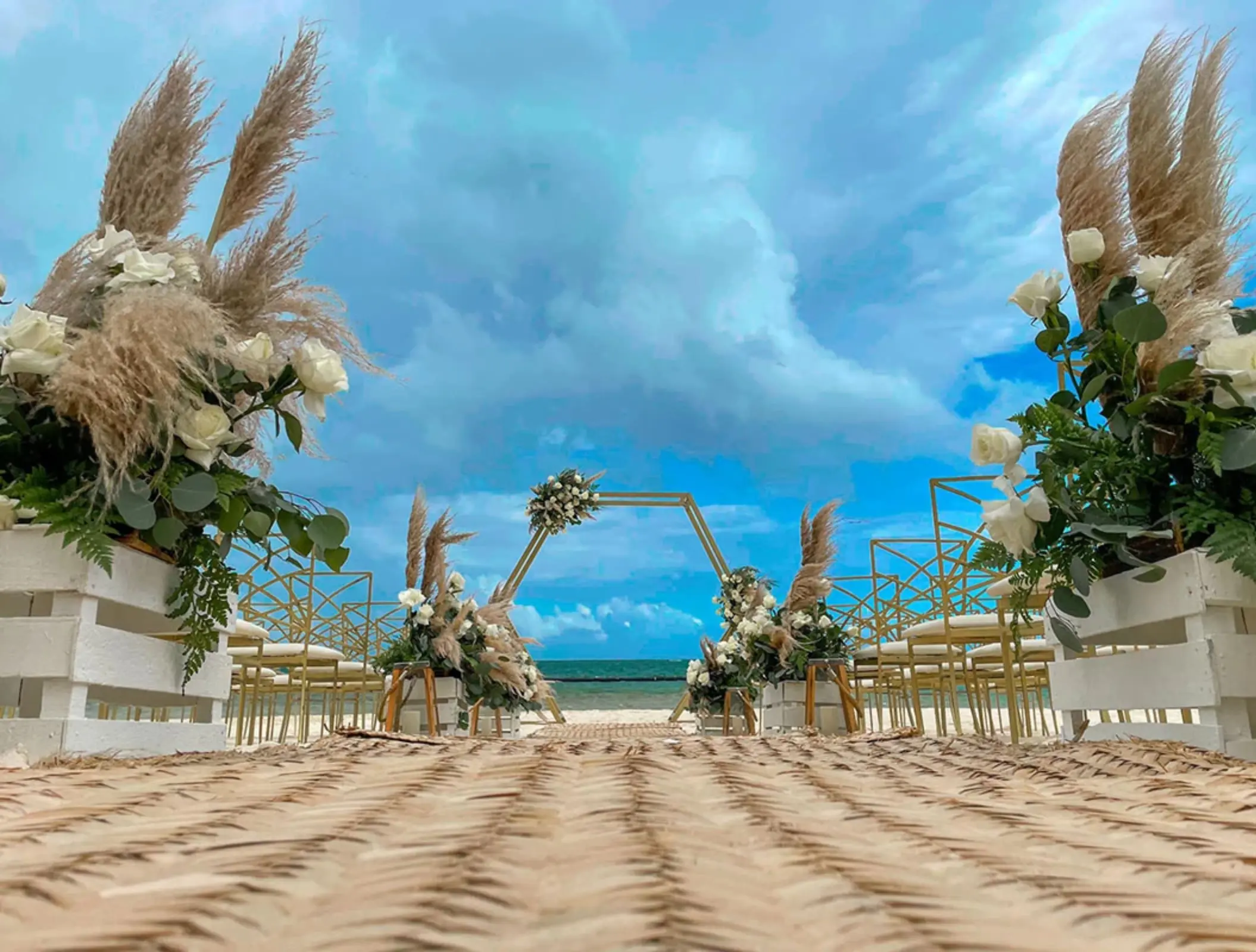 Romantic beach wedding setup with floral aisle, gold chairs, and hexagon arch.