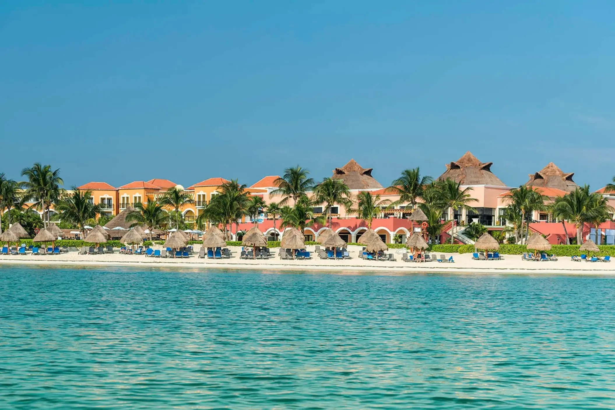 A wide shot of the resort's private beach with lounge chairs, palm trees, and the turquoise ocean.
