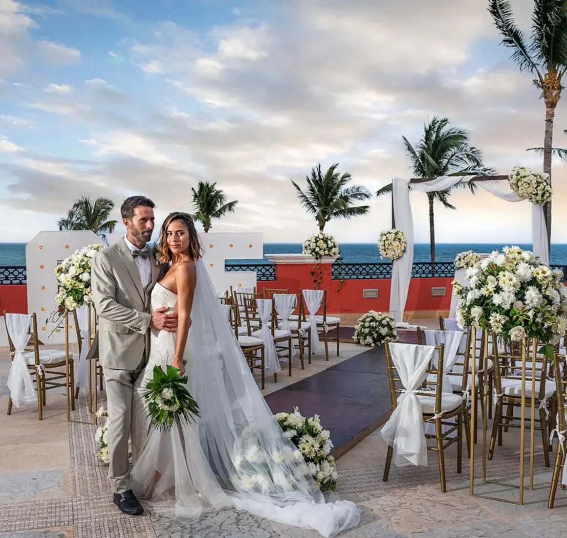 A bride and groom posing for a photo on a balcony with a wedding setup overlooking the ocean.