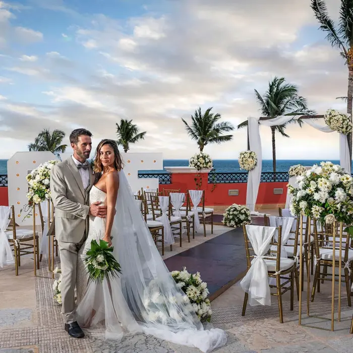 A bride and groom posing for a photo on a balcony with a wedding setup overlooking the ocean.