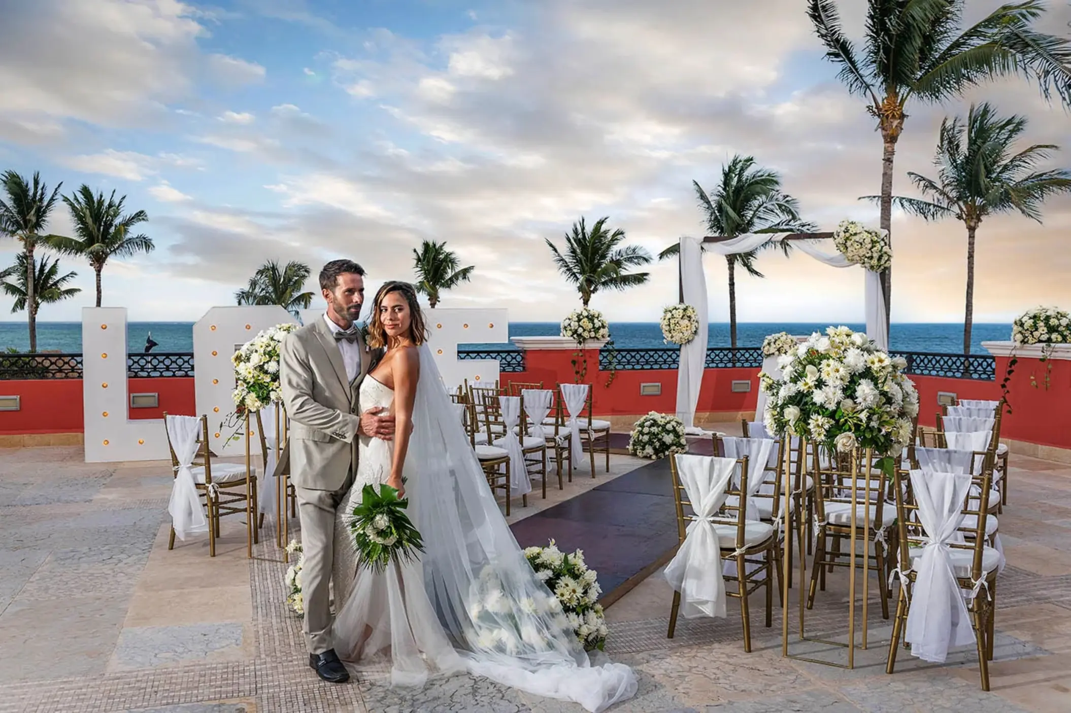 A bride and groom posing for a photo on a balcony with a wedding setup overlooking the ocean.
