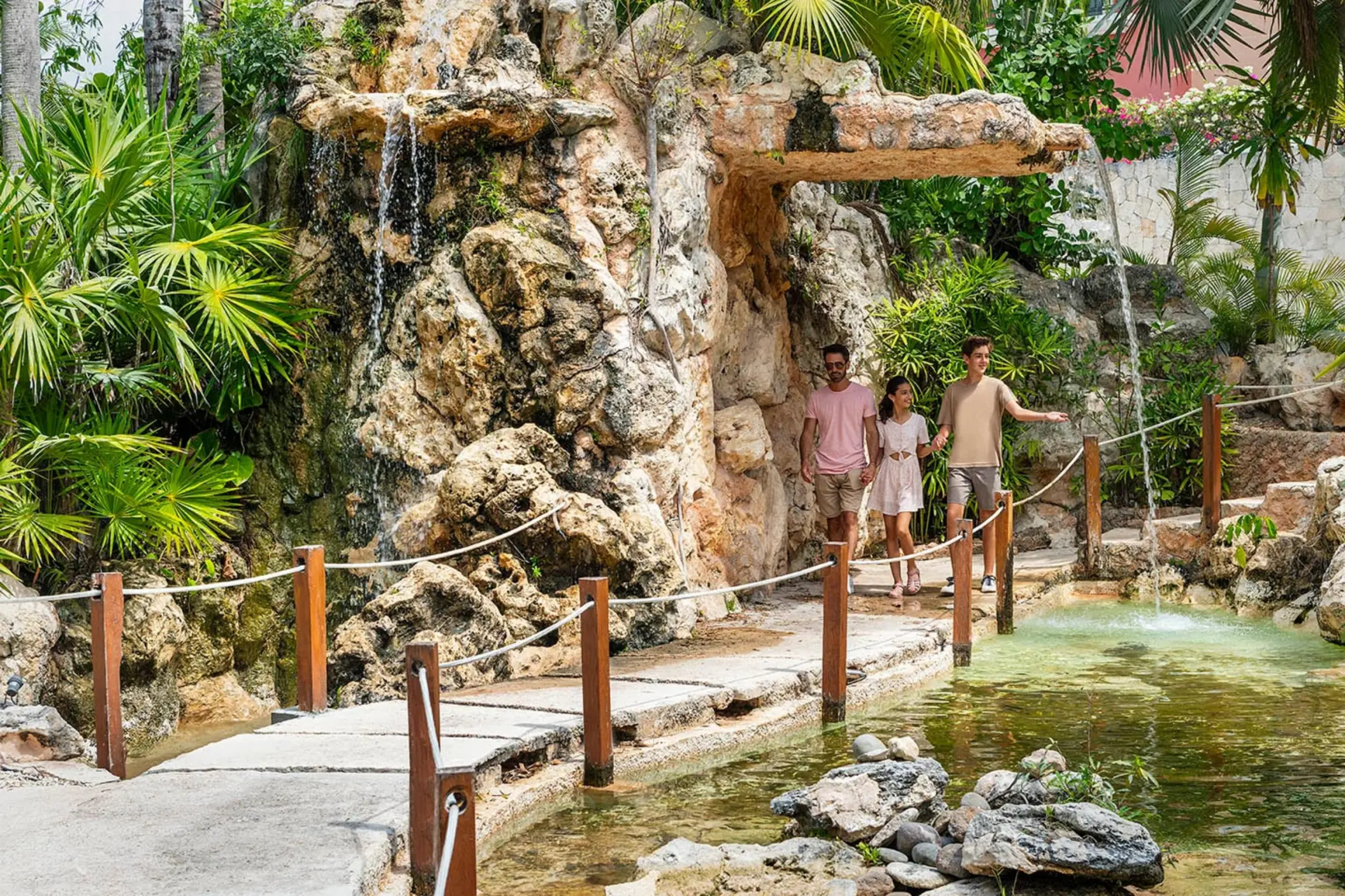 A family walking across a bridge over a pond, surrounded by lush gardens and a small waterfall.