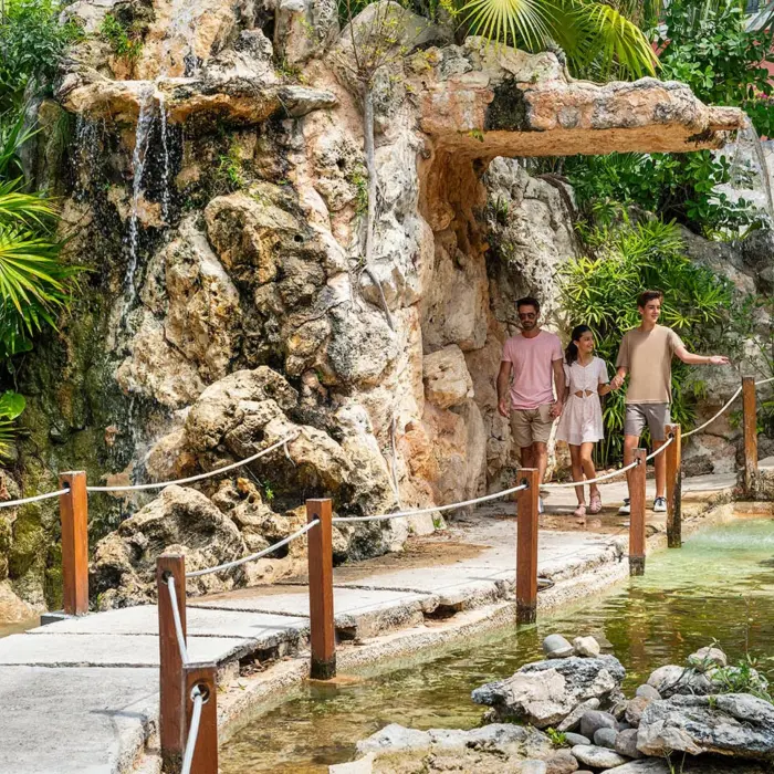 A family walking across a bridge over a pond, surrounded by lush gardens and a small waterfall.