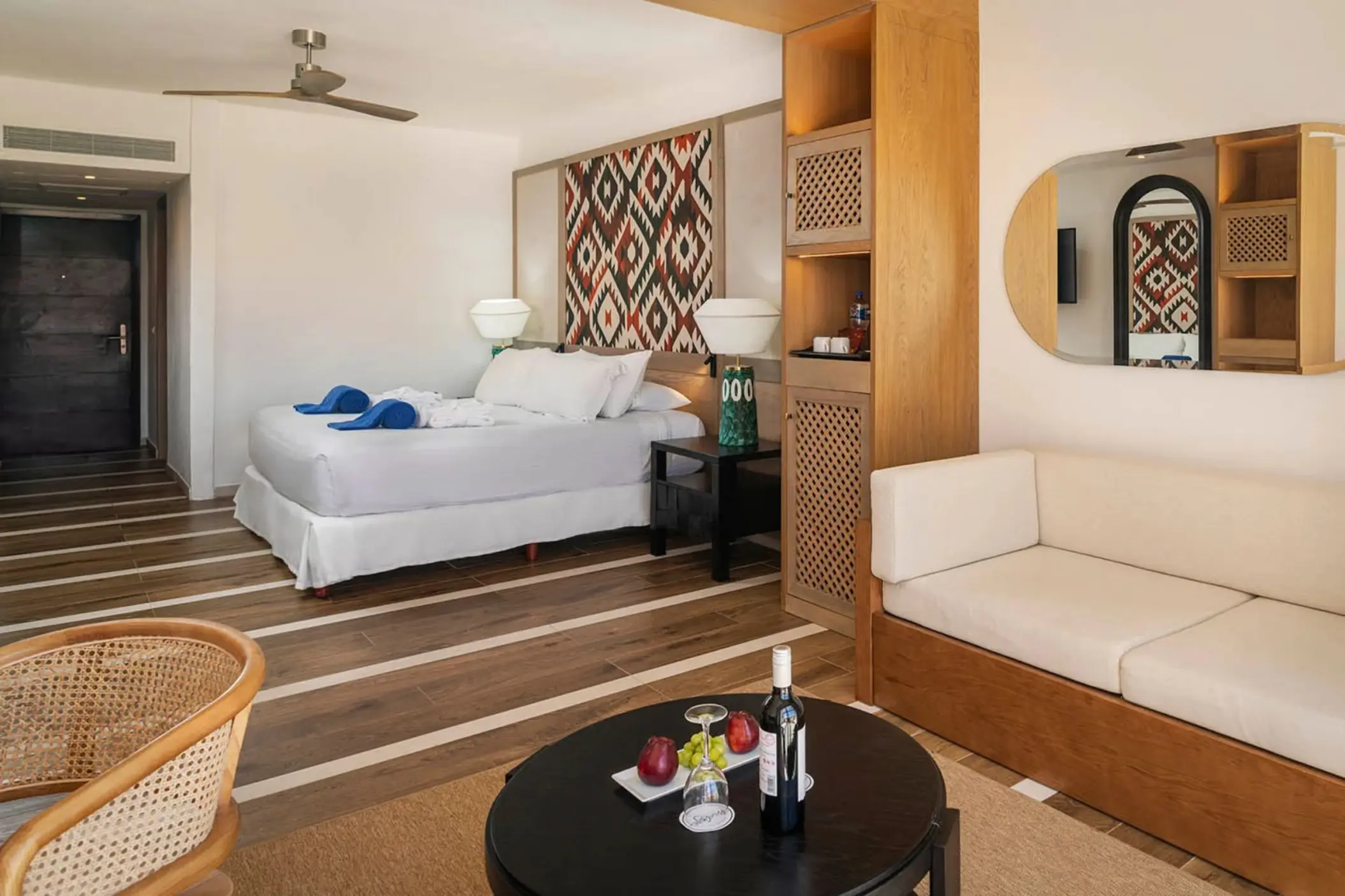 A modern suite with wood floors, a large bed, a couch, and a small table with wine and fruit.