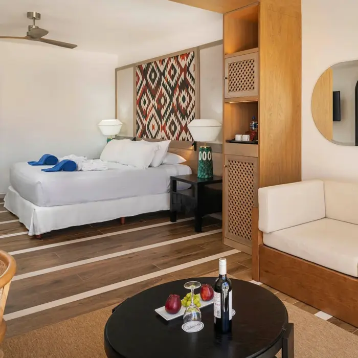 A modern suite with wood floors, a large bed, a couch, and a small table with wine and fruit.