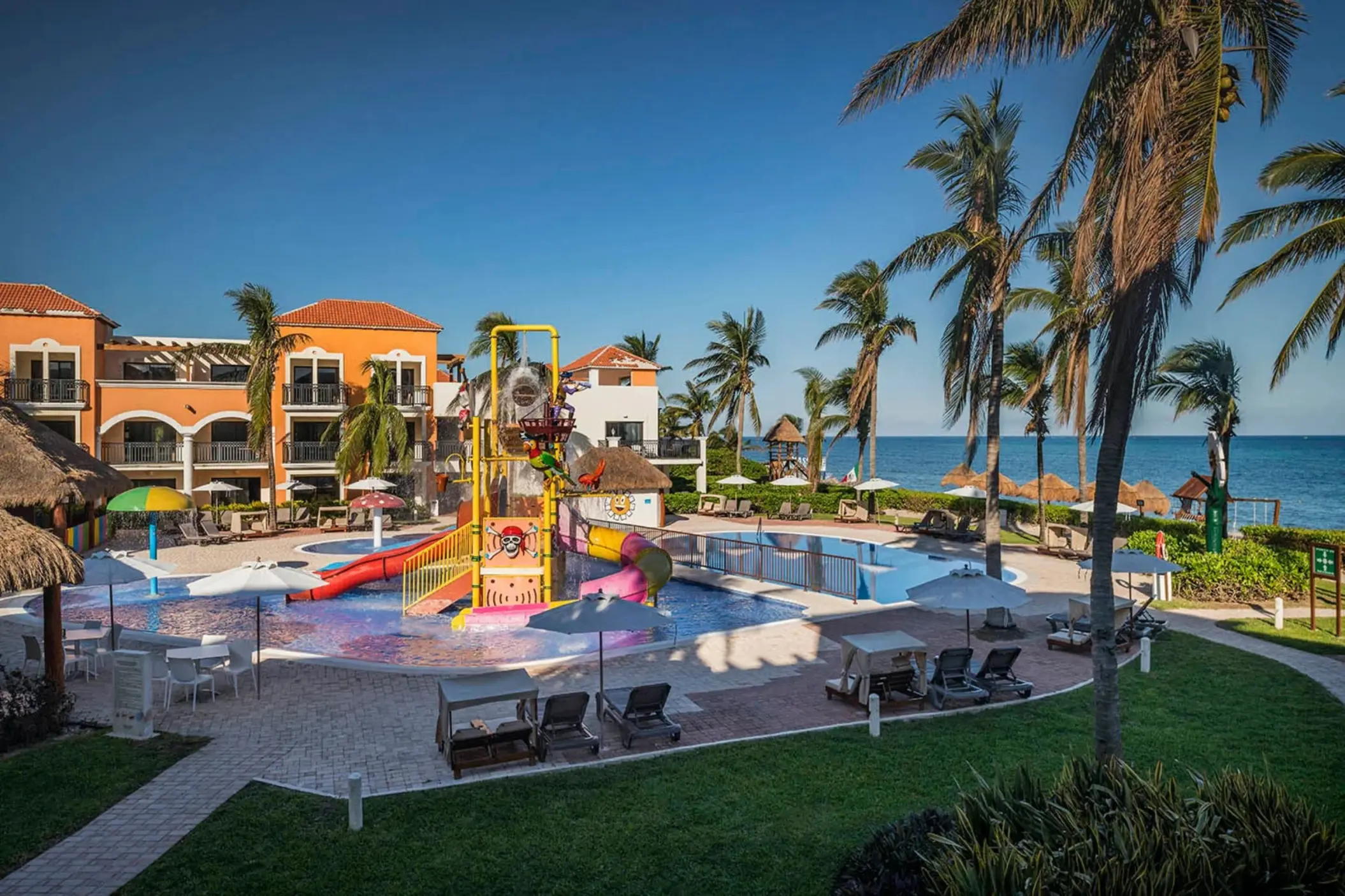 A vibrant kids' water park with a playground structure, slides, and pools, located by the ocean.