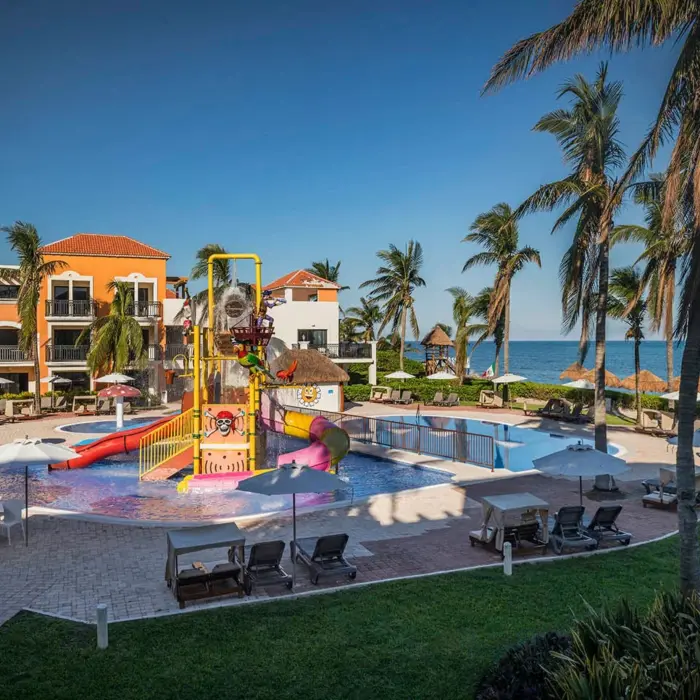 A vibrant kids' water park with a playground structure, slides, and pools, located by the ocean.