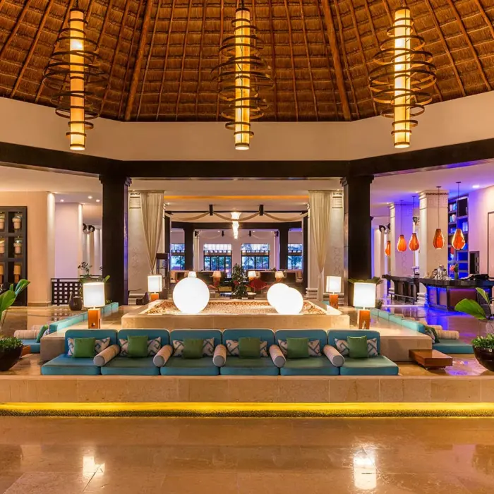 A spacious and luxurious hotel lobby with a thatched roof, large chandeliers, and a central seating area.