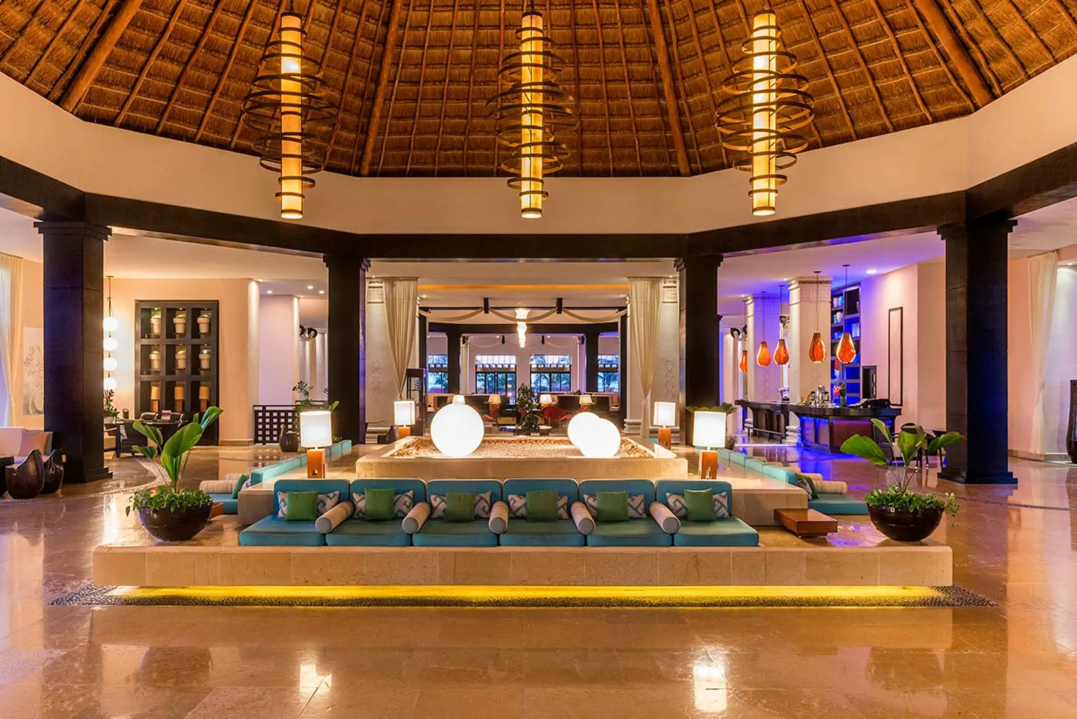 A spacious and luxurious hotel lobby with a thatched roof, large chandeliers, and a central seating area.