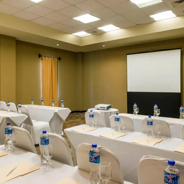 A meeting room with a projector screen and tables set up in a classroom style, with water bottles.