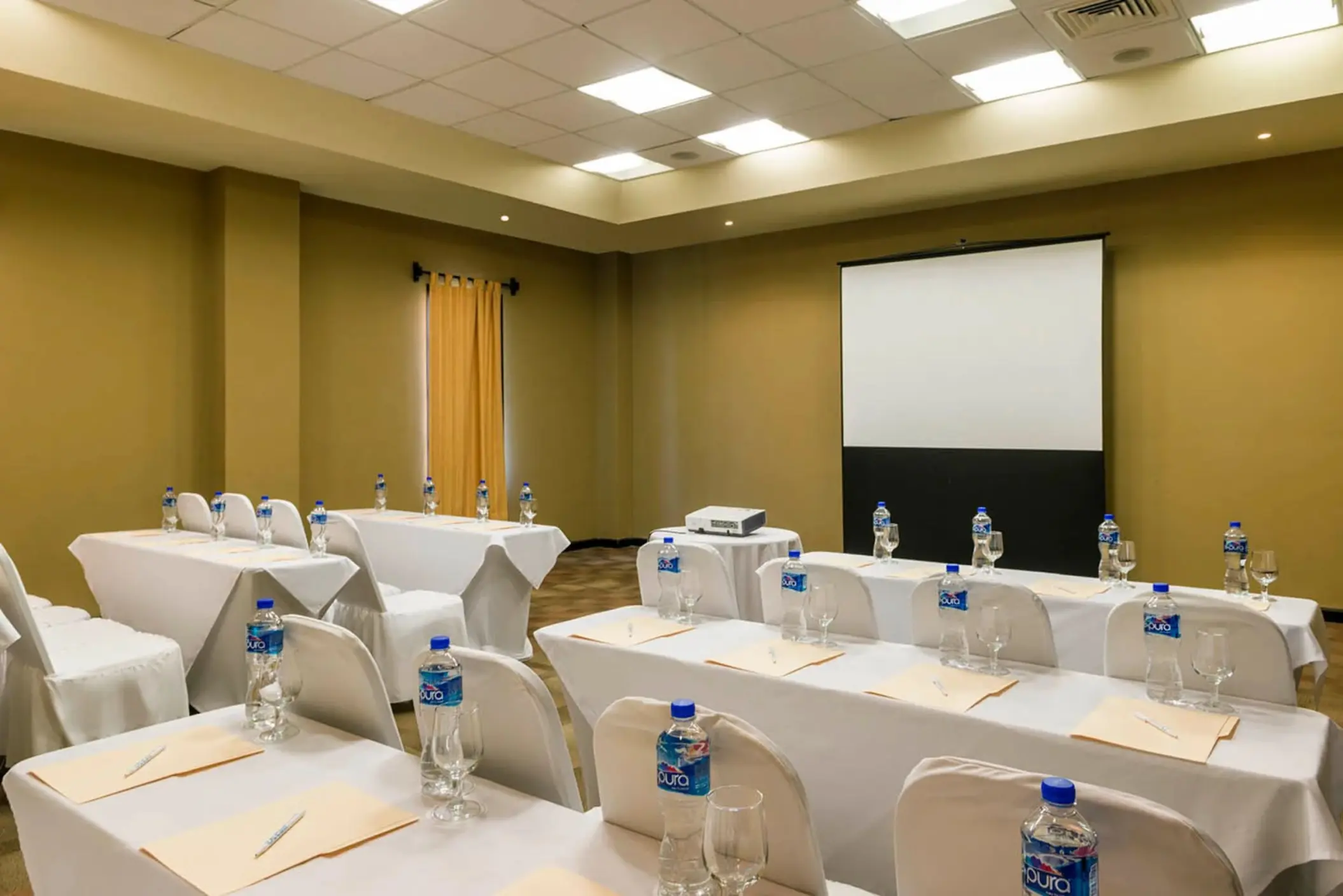 A meeting room with a projector screen and tables set up in a classroom style, with water bottles.