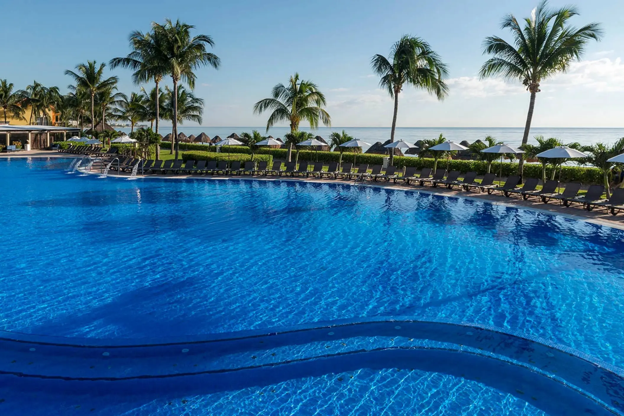 A large, sparkling blue pool with palm trees, lounge chairs, and the ocean visible in the background.