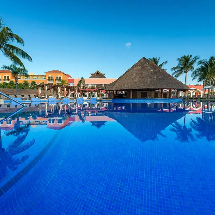 A view of a large swimming pool with a thatched-roof swim-up bar and resort buildings in the background.