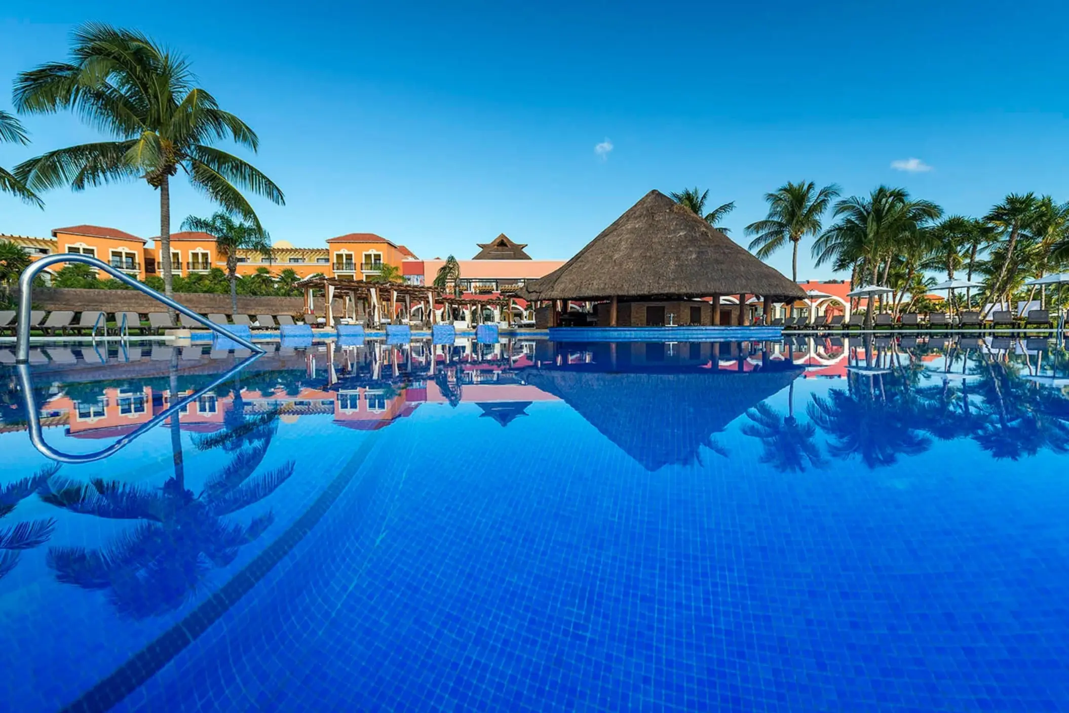 A view of a large swimming pool with a thatched-roof swim-up bar and resort buildings in the background.