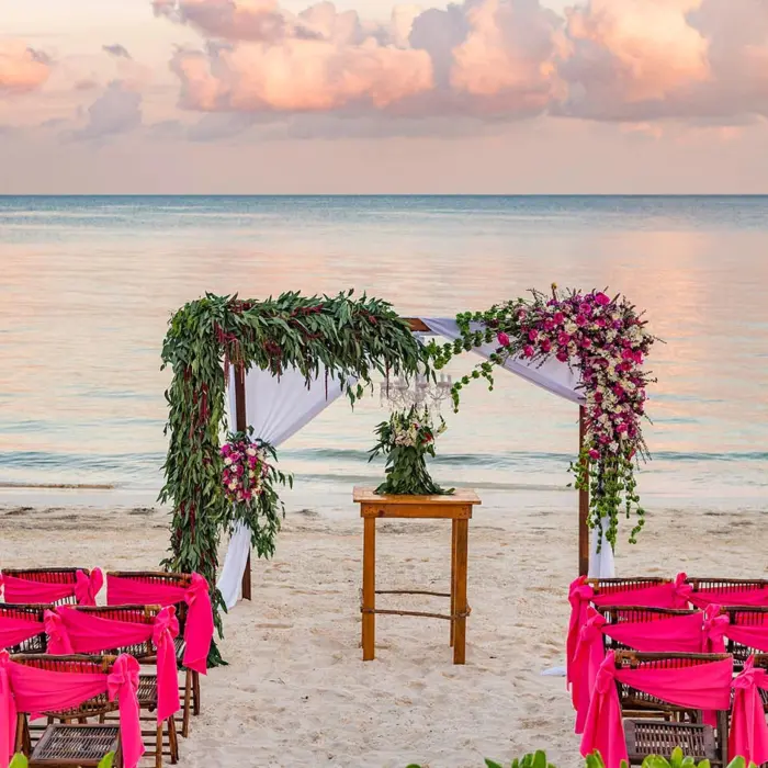A beach wedding setup with a floral arch and rows of chairs with bright pink sashes on the sand.