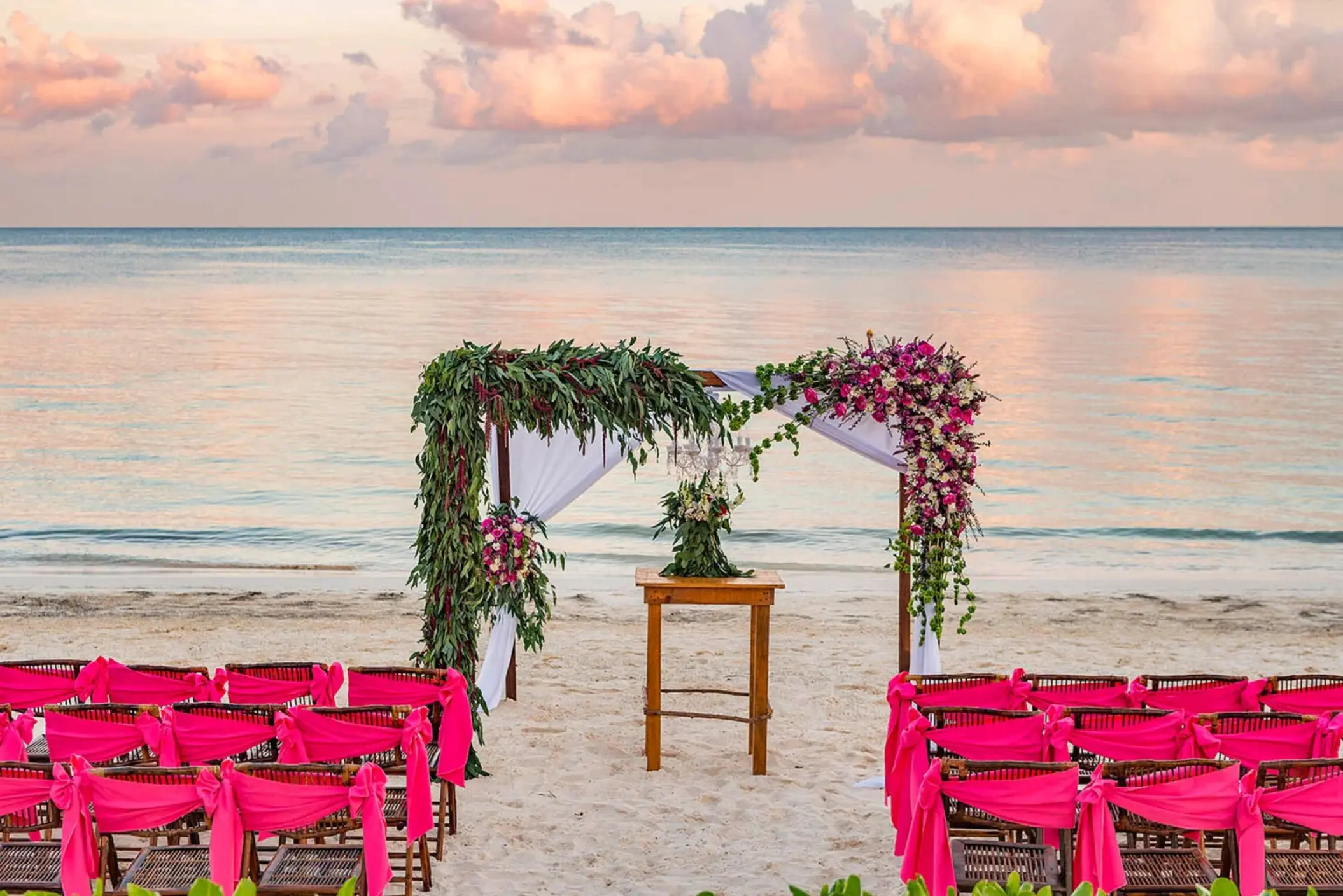 A beach wedding setup with a floral arch and rows of chairs with bright pink sashes on the sand.
