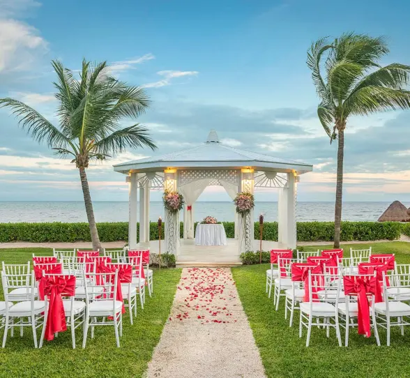 A beautiful outdoor wedding gazebo on a lawn with rows of chairs decorated with red bows, overlooking the ocean.