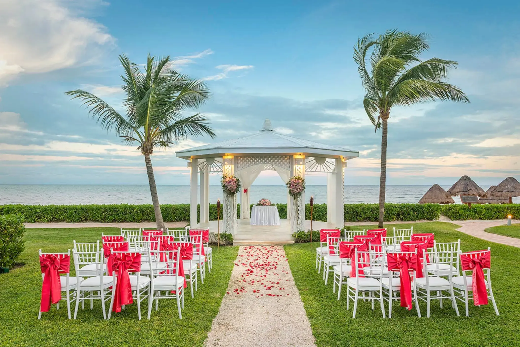 A beautiful outdoor wedding gazebo on a lawn with rows of chairs decorated with red bows, overlooking the ocean.