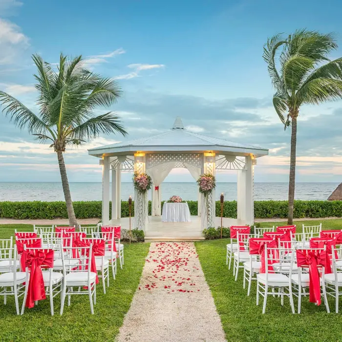 A beautiful outdoor wedding gazebo on a lawn with rows of chairs decorated with red bows, overlooking the ocean.