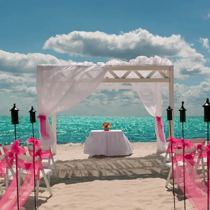 A romantic beach wedding setup with a white arch draped in sheer fabric, rows of chairs with pink sashes, and tiki torches by the turquoise ocean.