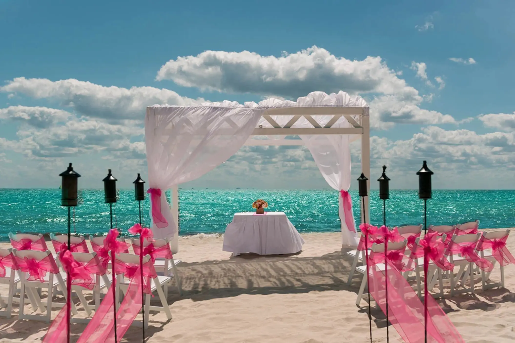 A romantic beach wedding setup with a white arch draped in sheer fabric, rows of chairs with pink sashes, and tiki torches by the turquoise ocean.