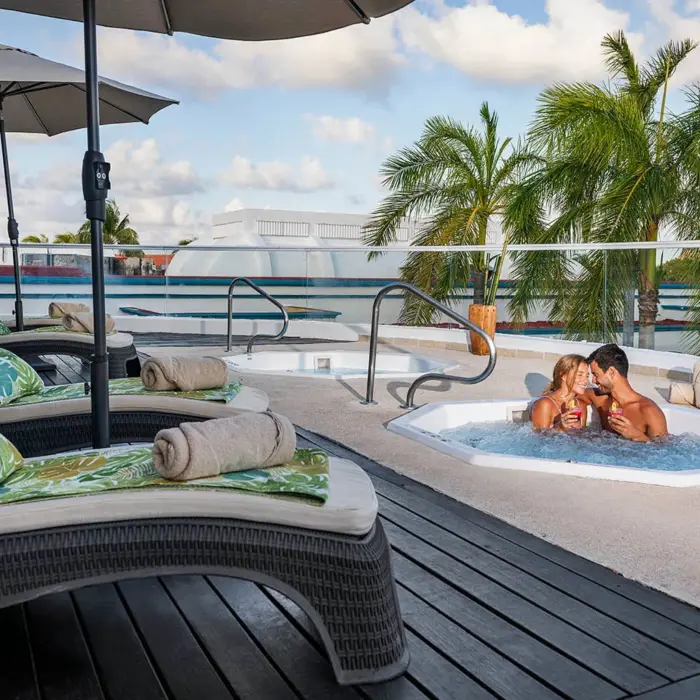 couple enjoying a bubbly hot tub on a rooftop terrace, surrounded by palm trees and lounge chairs under a clear sky.