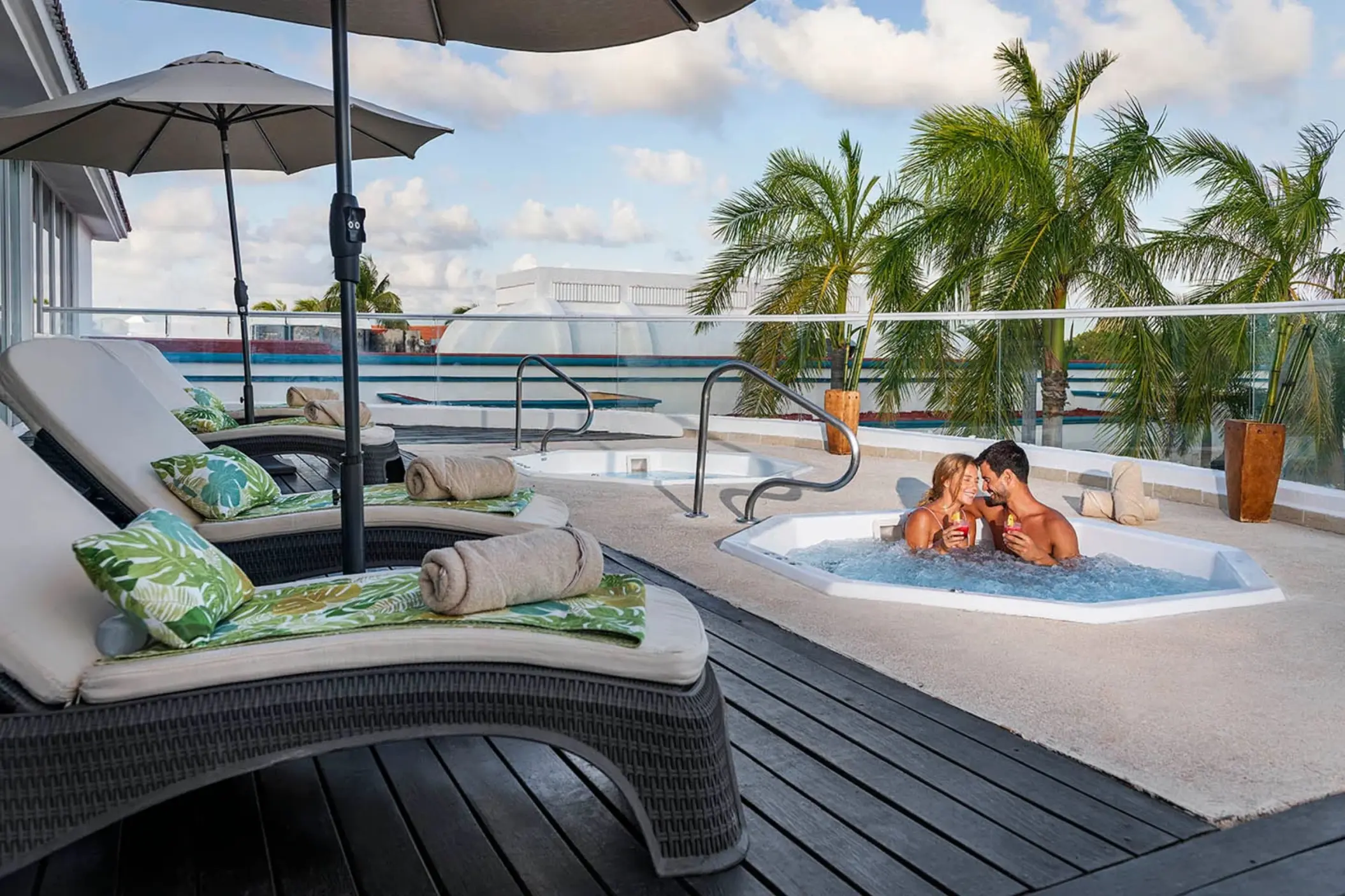 couple enjoying a bubbly hot tub on a rooftop terrace, surrounded by palm trees and lounge chairs under a clear sky.