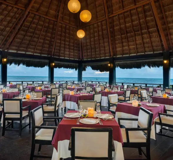 elegant open-air restaurant with a high thatched roof, formal dining tables with white and red settings, and an expansive ocean view.