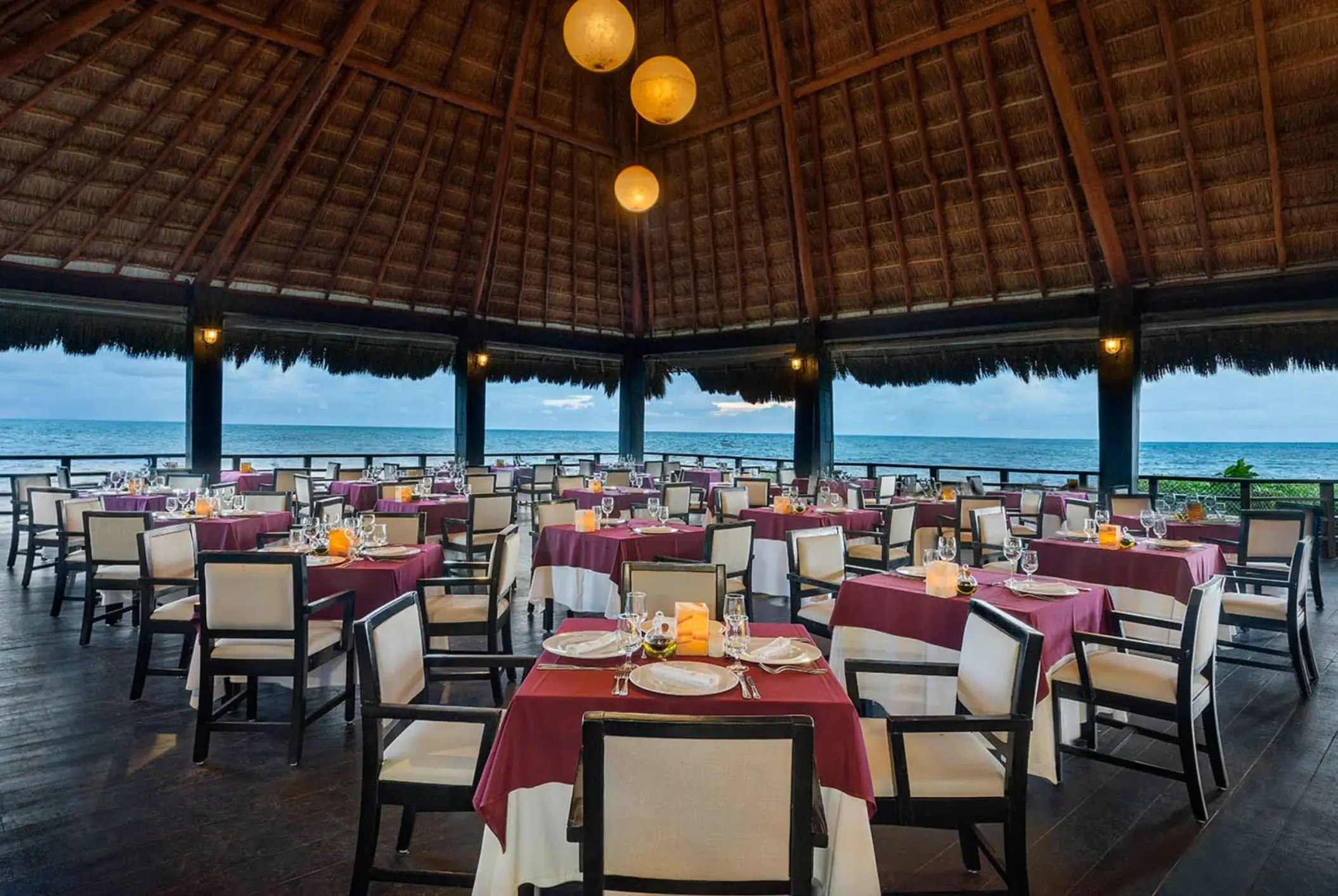 elegant open-air restaurant with a high thatched roof, formal dining tables with white and red settings, and an expansive ocean view.
