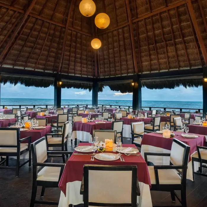 elegant open-air restaurant with a high thatched roof, formal dining tables with white and red settings, and an expansive ocean view.