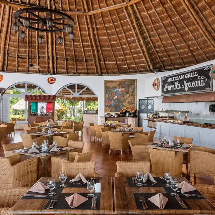 A rustic Mexican grill restaurant with a high thatched roof, wicker chairs, and a buffet area, offering an open-air dining experience.