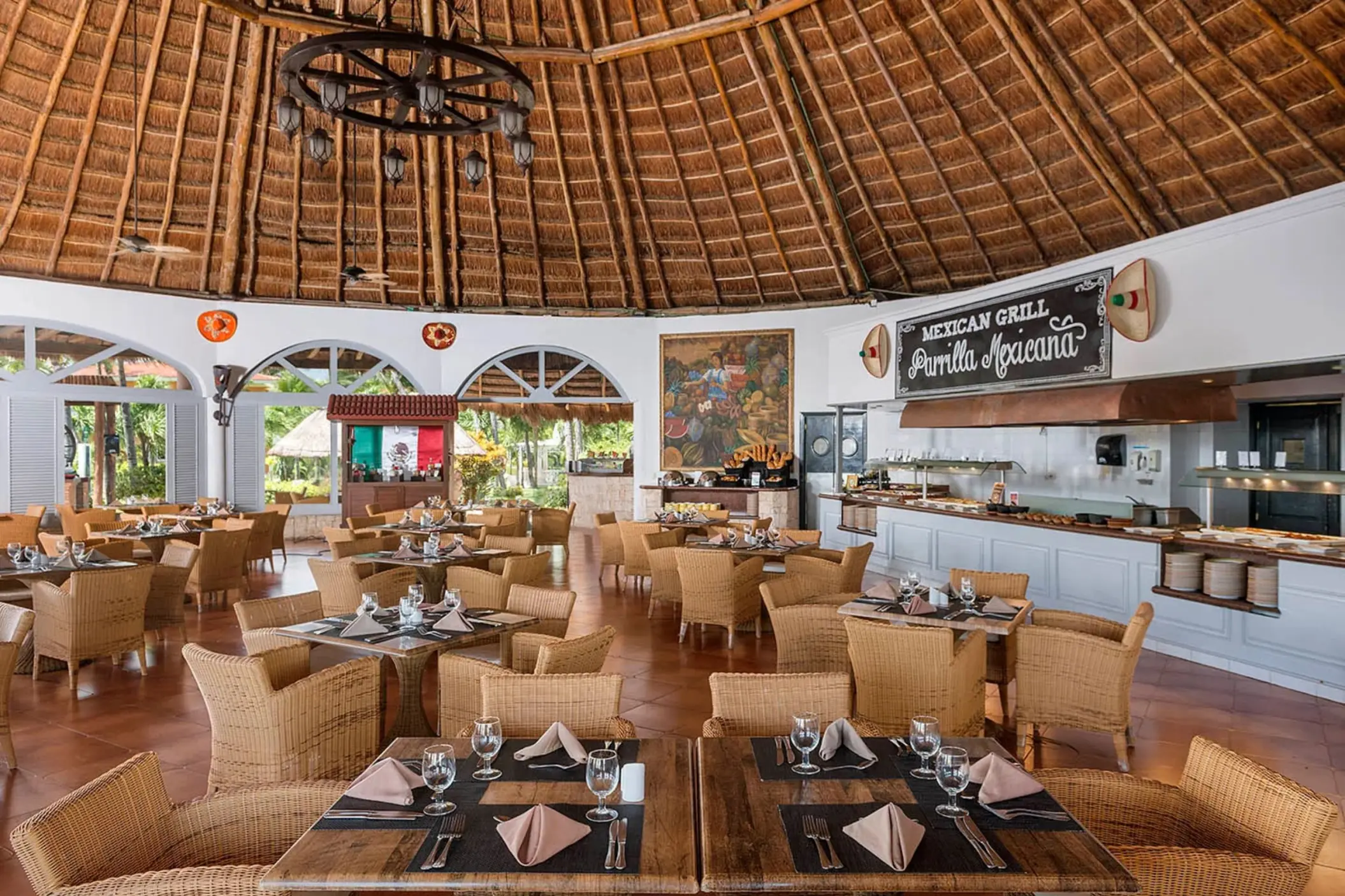 A rustic Mexican grill restaurant with a high thatched roof, wicker chairs, and a buffet area, offering an open-air dining experience.