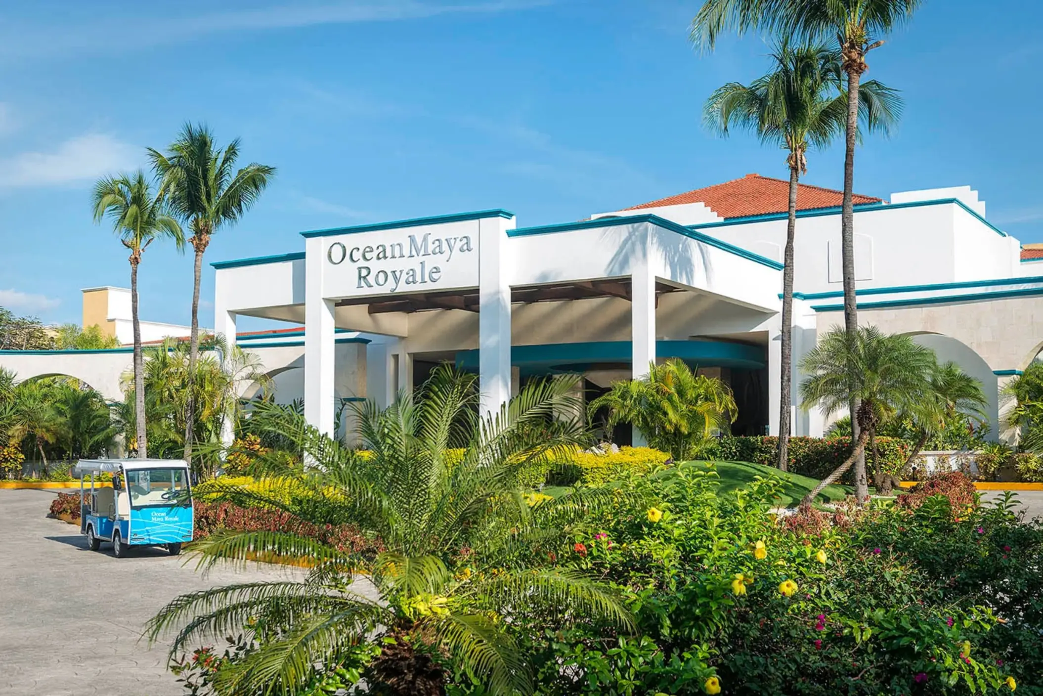 The main entrance of Ocean Maya Royale resort, featuring a grand white building, lush tropical landscaping, and a blue golf cart.