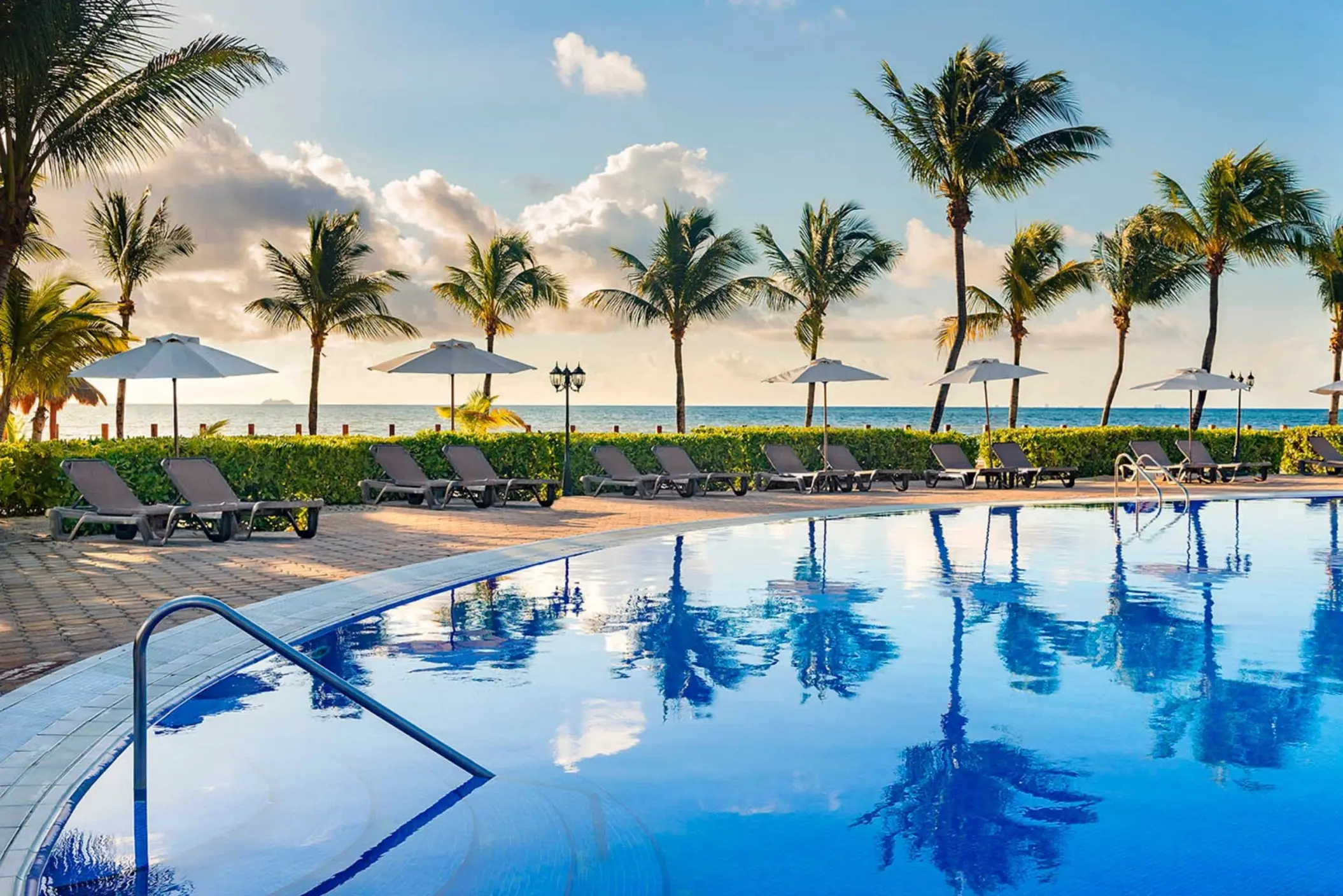 stunning outdoor pool with numerous lounge chairs, umbrellas, and tall palm trees, facing the ocean under a beautiful sky.