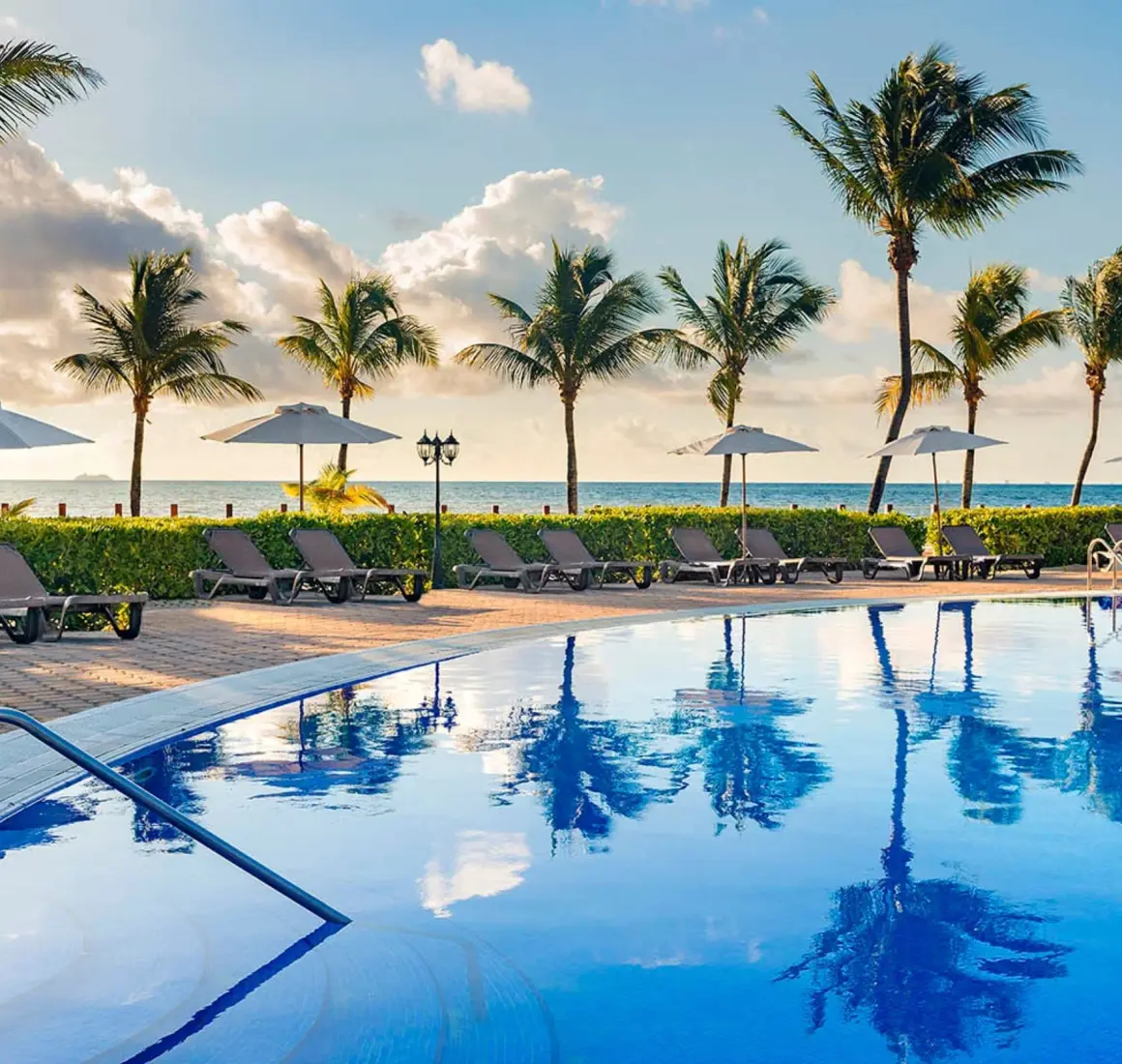 stunning outdoor pool with numerous lounge chairs, umbrellas, and tall palm trees, facing the ocean under a beautiful sky.