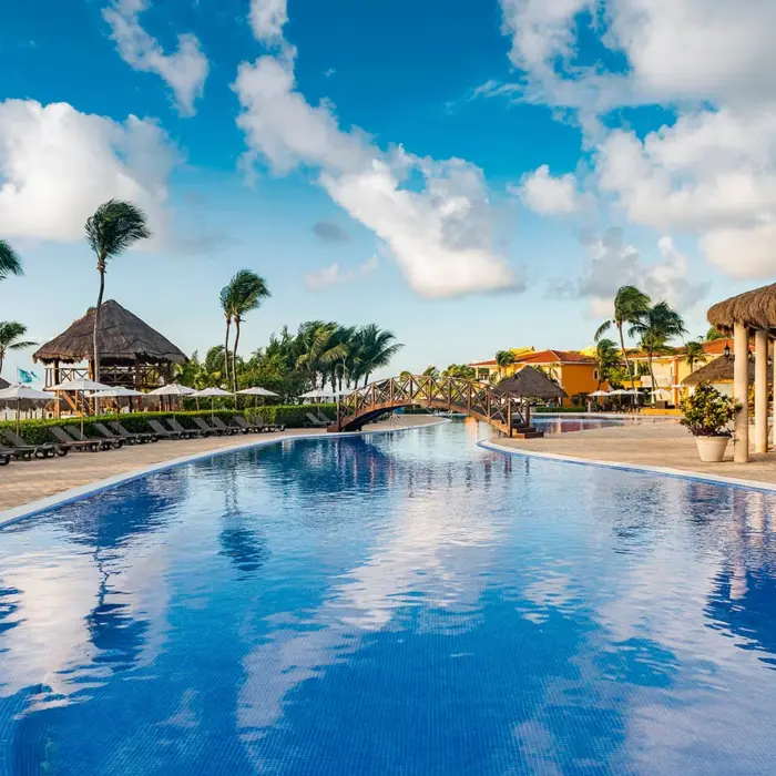 winding resort pool with a charming wooden bridge, surrounded by palm trees, lounge chairs, and resort buildings under a blue sky.