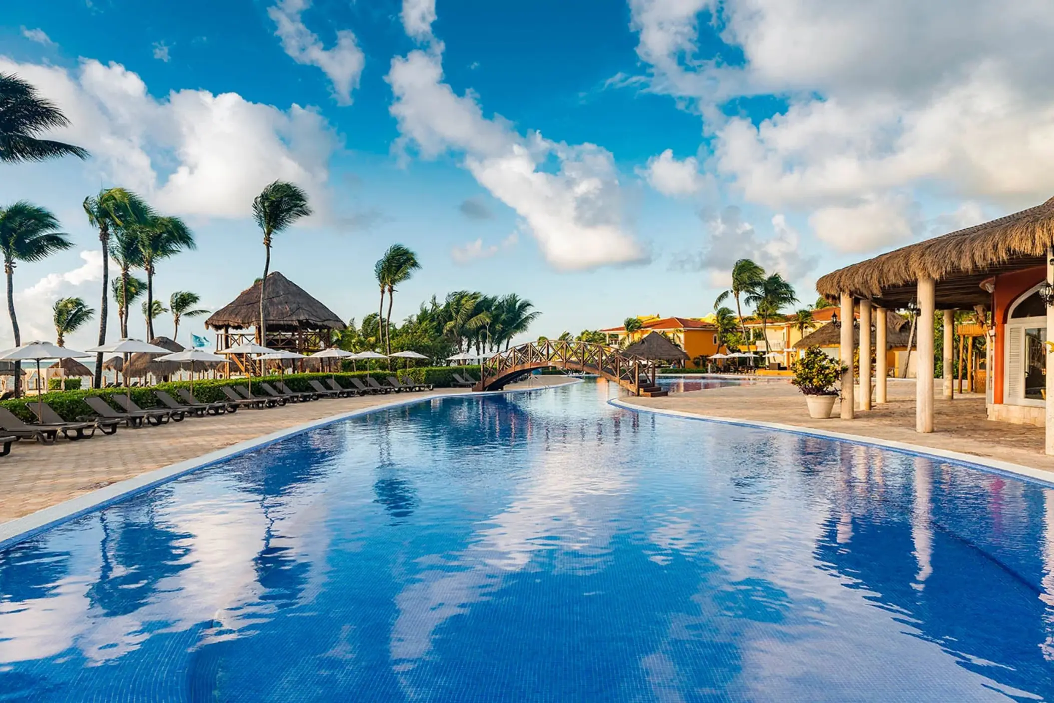 winding resort pool with a charming wooden bridge, surrounded by palm trees, lounge chairs, and resort buildings under a blue sky.