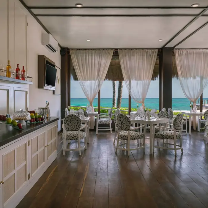An elegant lounge with an ocean view, featuring white and patterned chairs, dark wood floors, and a buffet counter with drinks.
