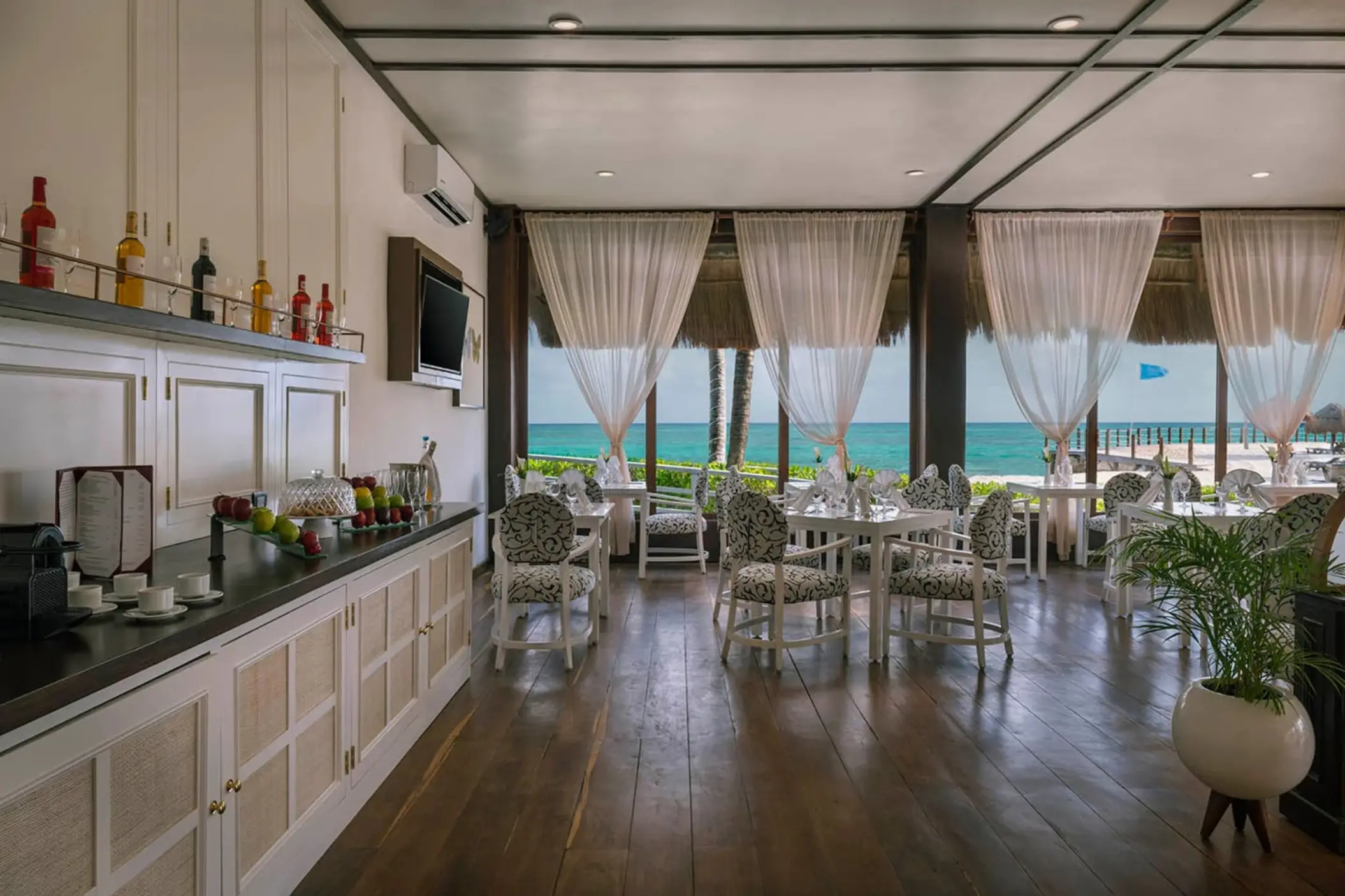 An elegant lounge with an ocean view, featuring white and patterned chairs, dark wood floors, and a buffet counter with drinks.