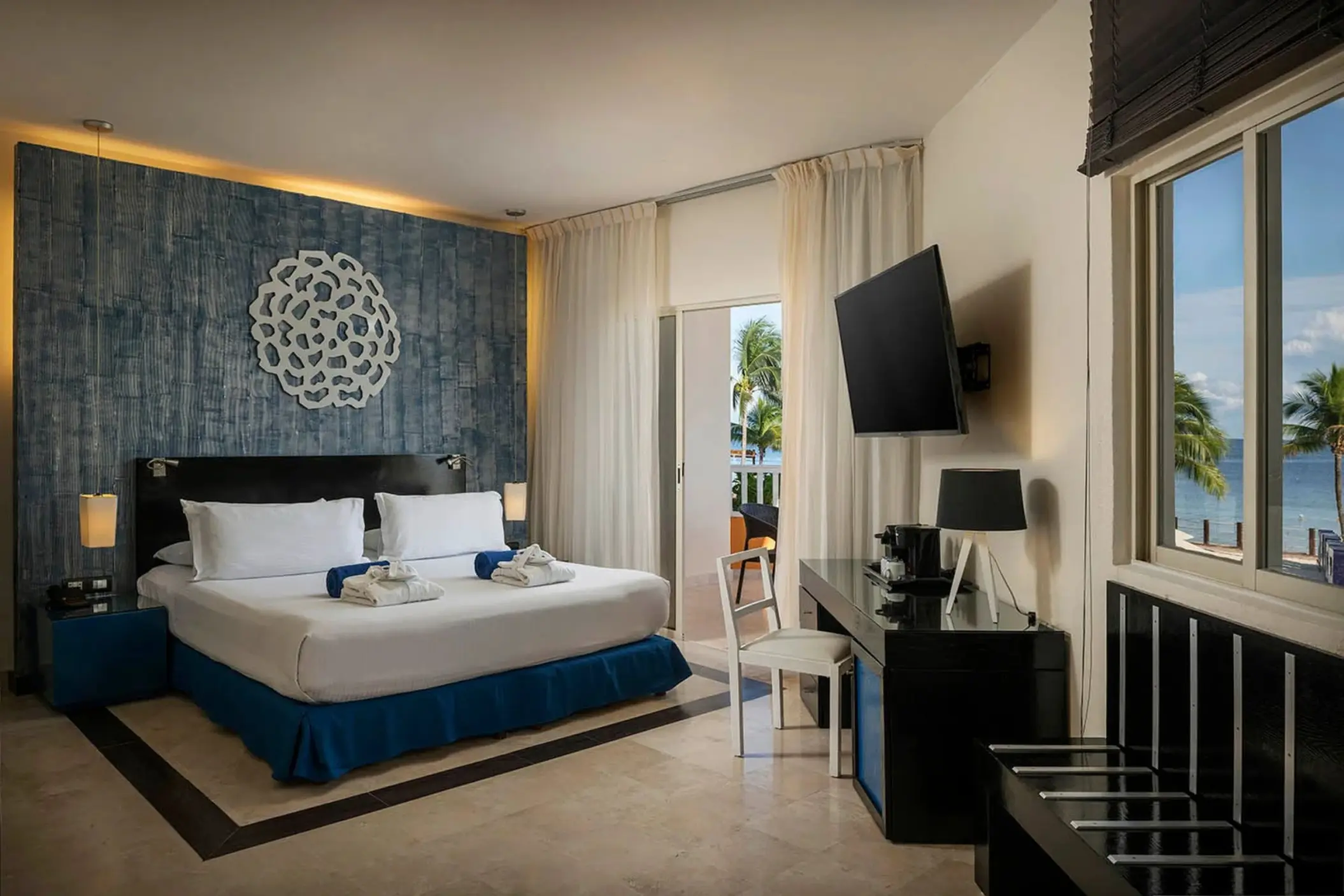 bright oceanfront suite with a king bed, blue patterned wall, wooden blinds, and a balcony overlooking the ocean.