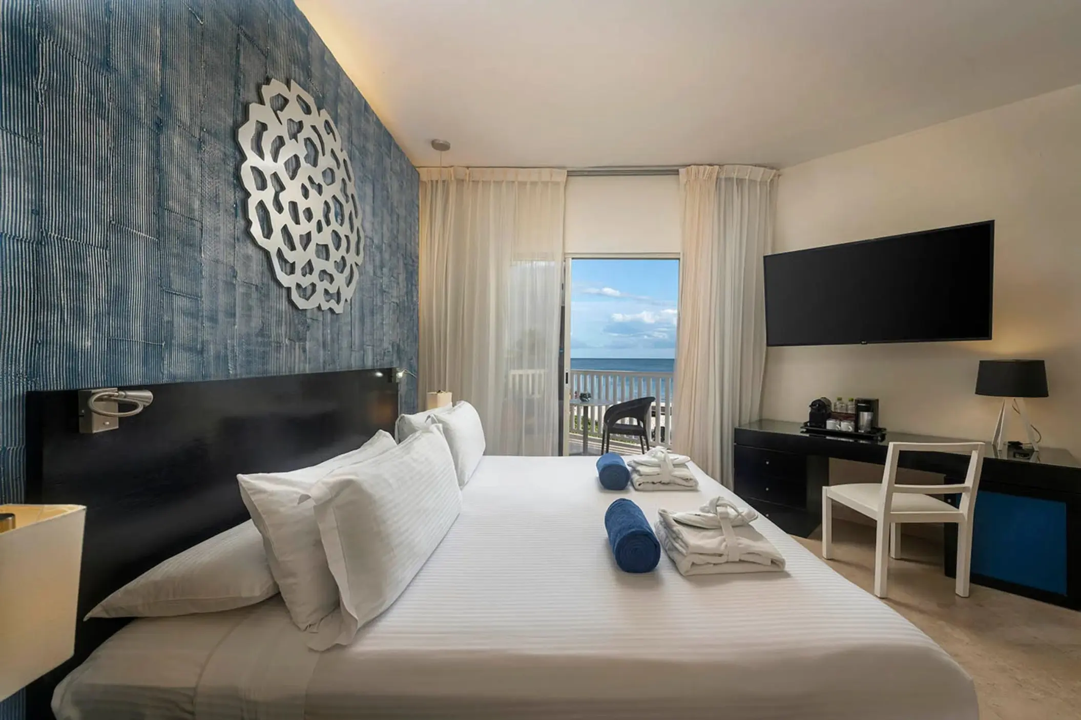 modern Privilege Maya Deluxe Ocean Front suite with a king bed, ocean view balcony, blue accent wall, and flat-screen TV.
