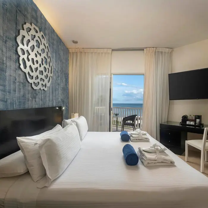 modern Privilege Maya Deluxe Ocean Front suite with a king bed, ocean view balcony, blue accent wall, and flat-screen TV.