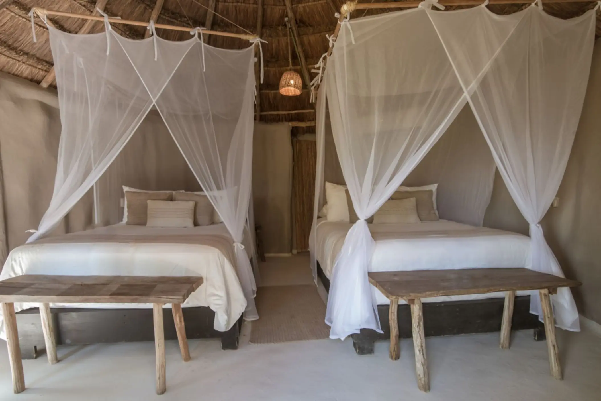 Interior of a rustic cabana with a thatched roof, featuring two beds draped with mosquito nets and simple wooden furniture.