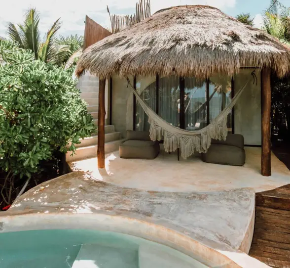 A charming casita with a thatched roof, hammock, small plunge pool, and lush greenery, offering an ocean view.
